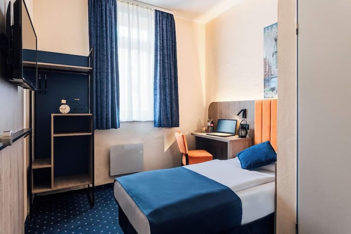 Hotellihuone, jossa on sänky, työpöytä tietokoneella, tuoli, hylly, jossa on kello, ja ikkuna, jossa on verhot.
