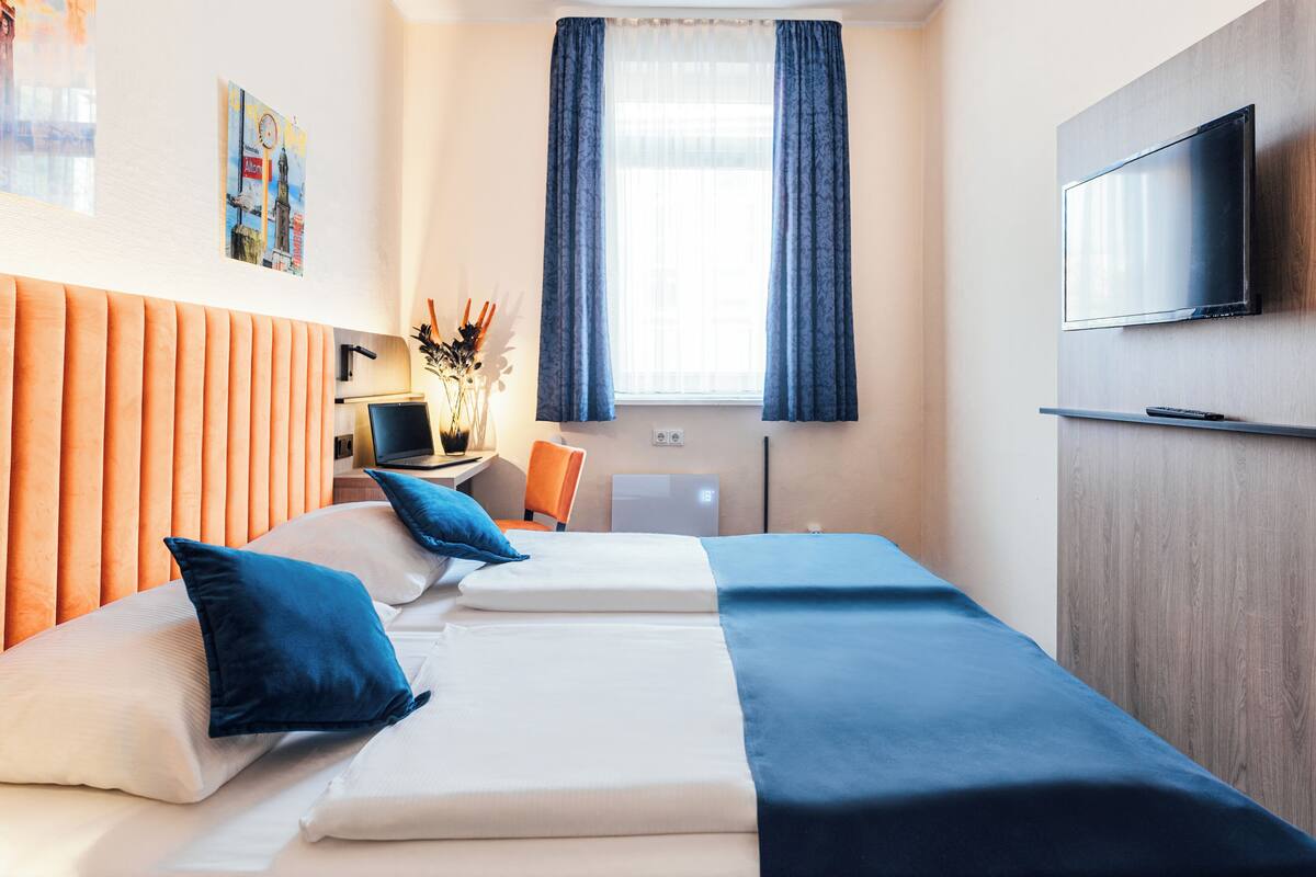 Hotellihuone, jossa on sänky, työpöytä kannettavalla tietokoneella, televisio ja ikkuna, jossa on verhot.