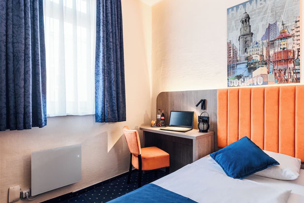 Hotellihuone, jossa on sänky, työpöytä, tietokone, tuoli ja patteri.