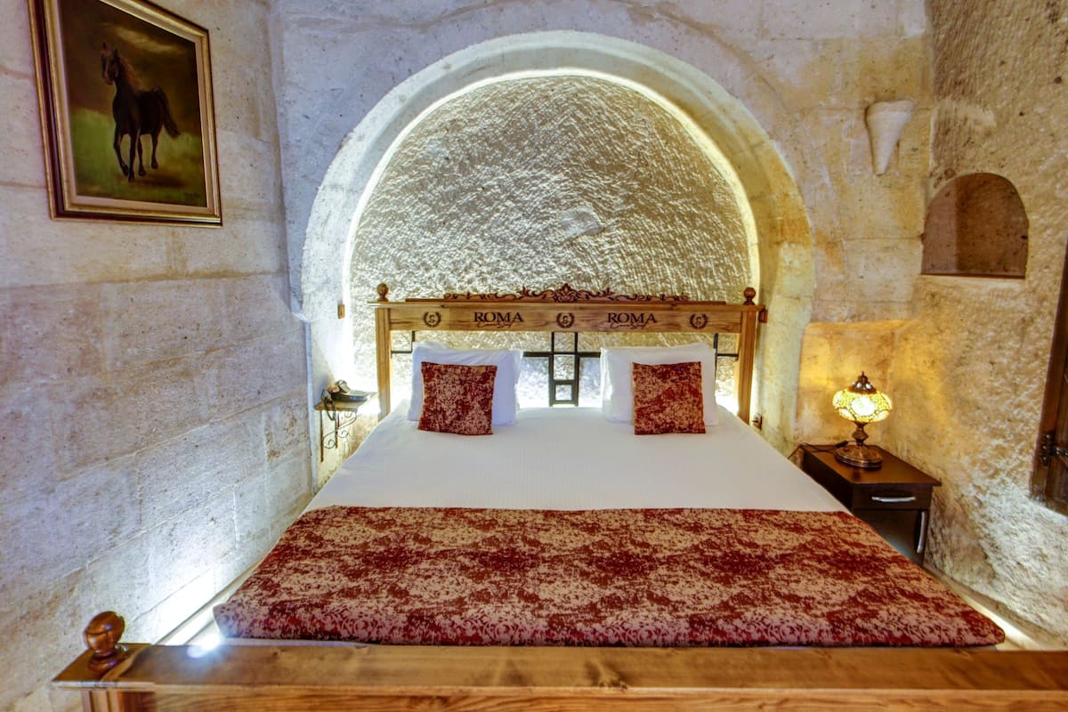 Un dormitorio con una cama grande, cabecera de madera, colcha estampada en rojo y blanco, un cuadro enmarcado en la pared y una pared de piedra con un ventanal de arco.