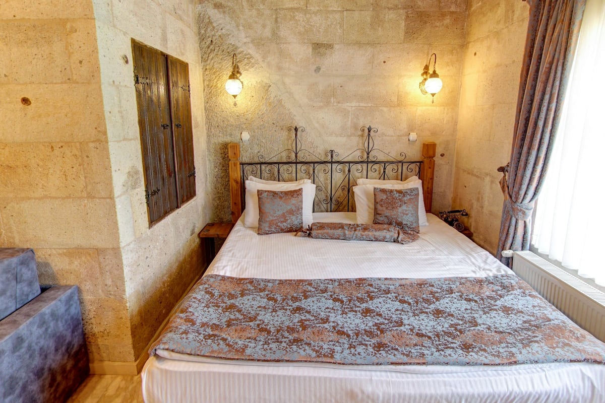 Un dormitorio con techo de madera, paredes de piedra, cama con ropa de cama estampada, dos apliques de pared y una ventana con cortinas.