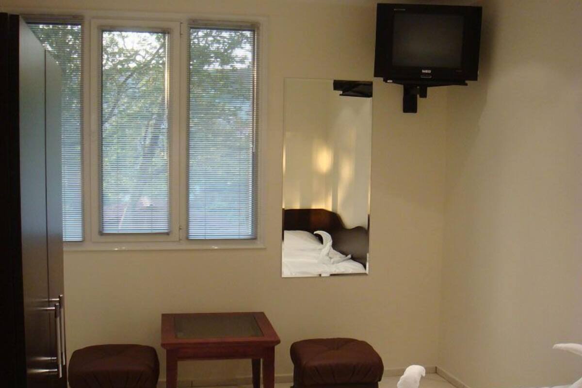 A hotel room with a bed, a small table, two armchairs, a television, and a window with blinds.