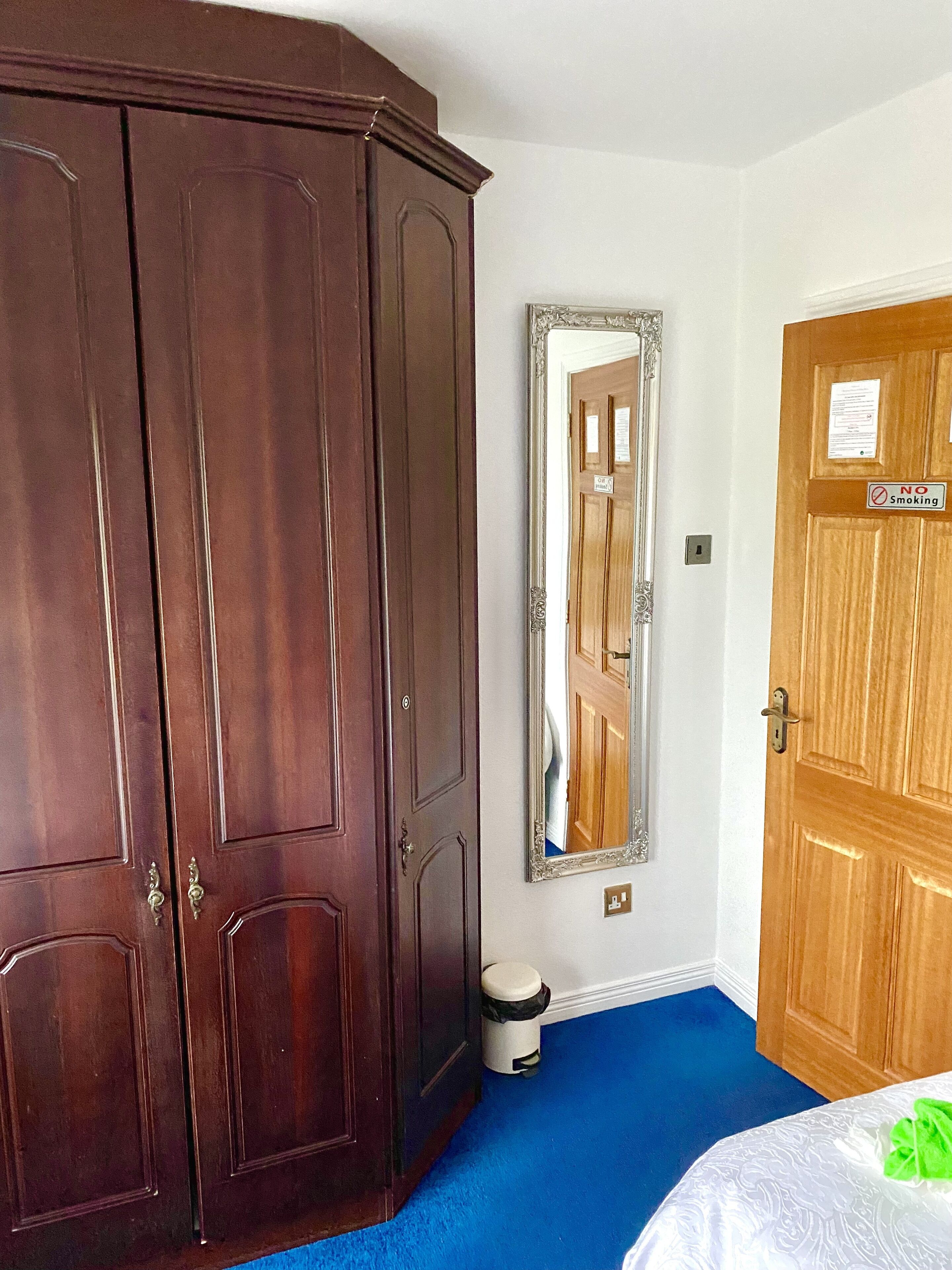 A room with a large wooden wardrobe, a mirror, a wooden door with a 'No Smoking' sign, and a blue carpet.