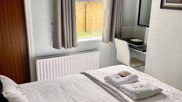 Una cama bien hecha con edredón blanco, una mesita de noche con lámpara, una ventana con cortinas y un cuadro en la pared.