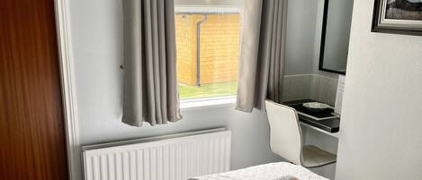 A neatly made bed with a white duvet, a small bedside table with a lamp, a window with curtains, and a framed picture on the wall.