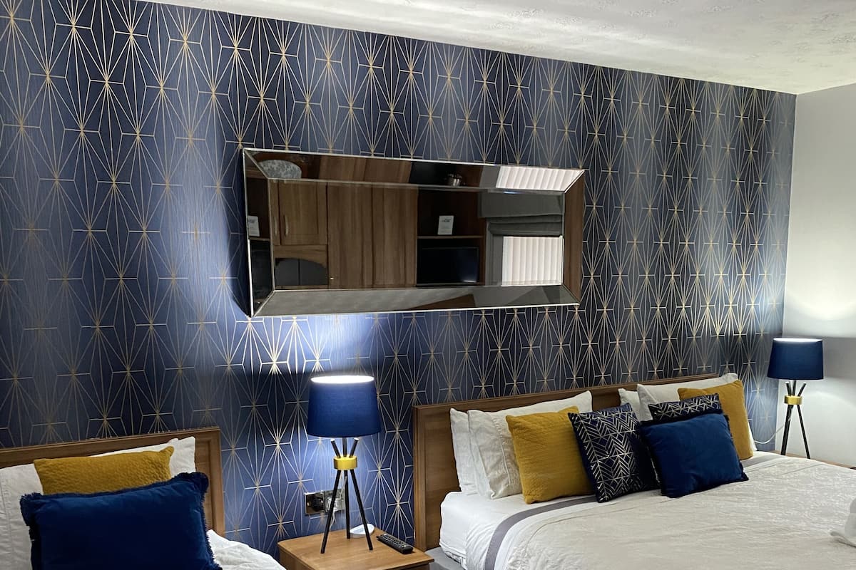 A hotel room with two beds, a nightstand with a lamp, a mirror above the bed, and a patterned wall.