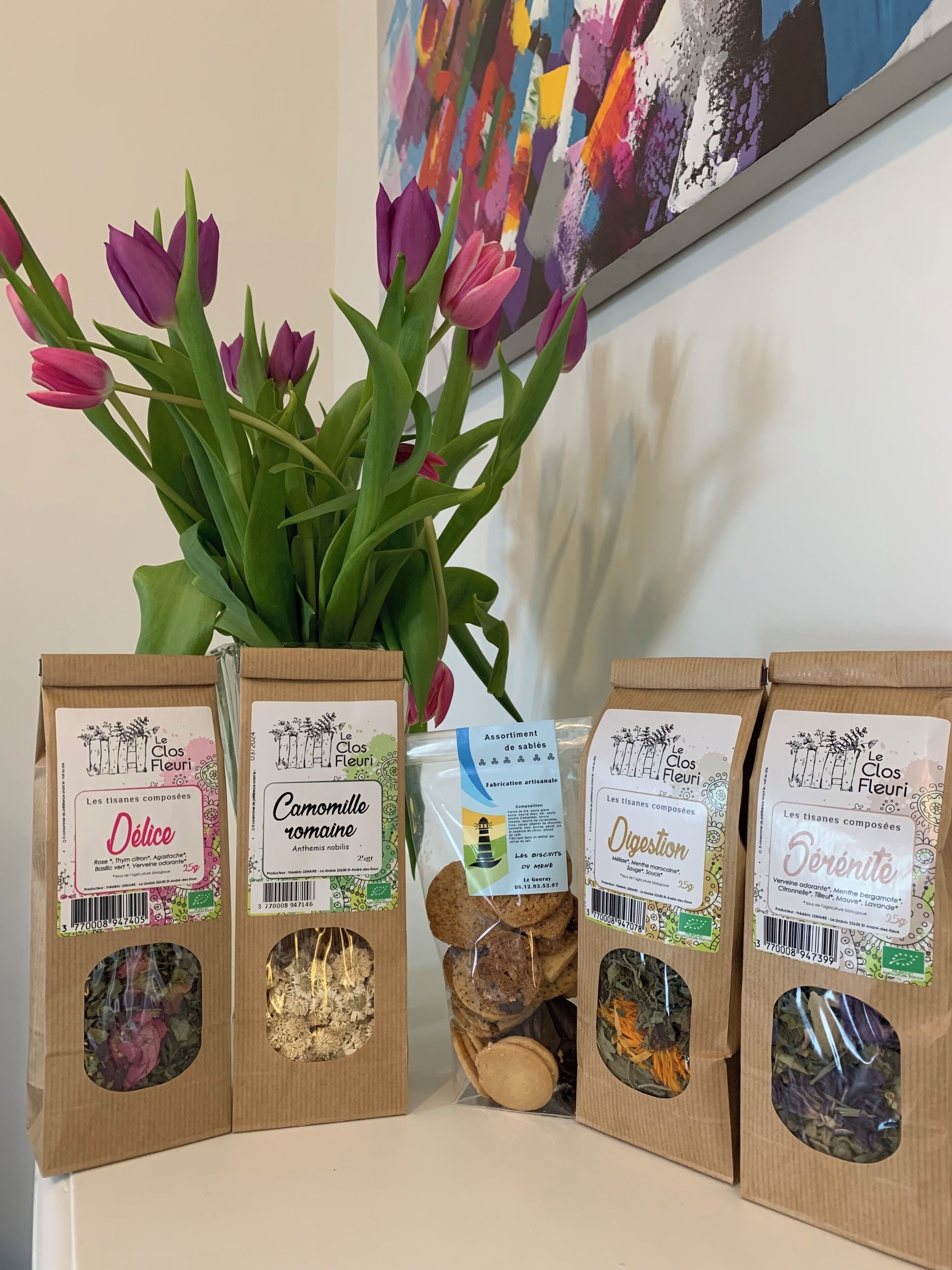 Quatre sacs en papier brun, chacun portant une étiquette et contenant différents produits à base de plantes, sont placés sur une étagère. En arrière-plan, on aperçoit un vase rempli de tulipes roses.