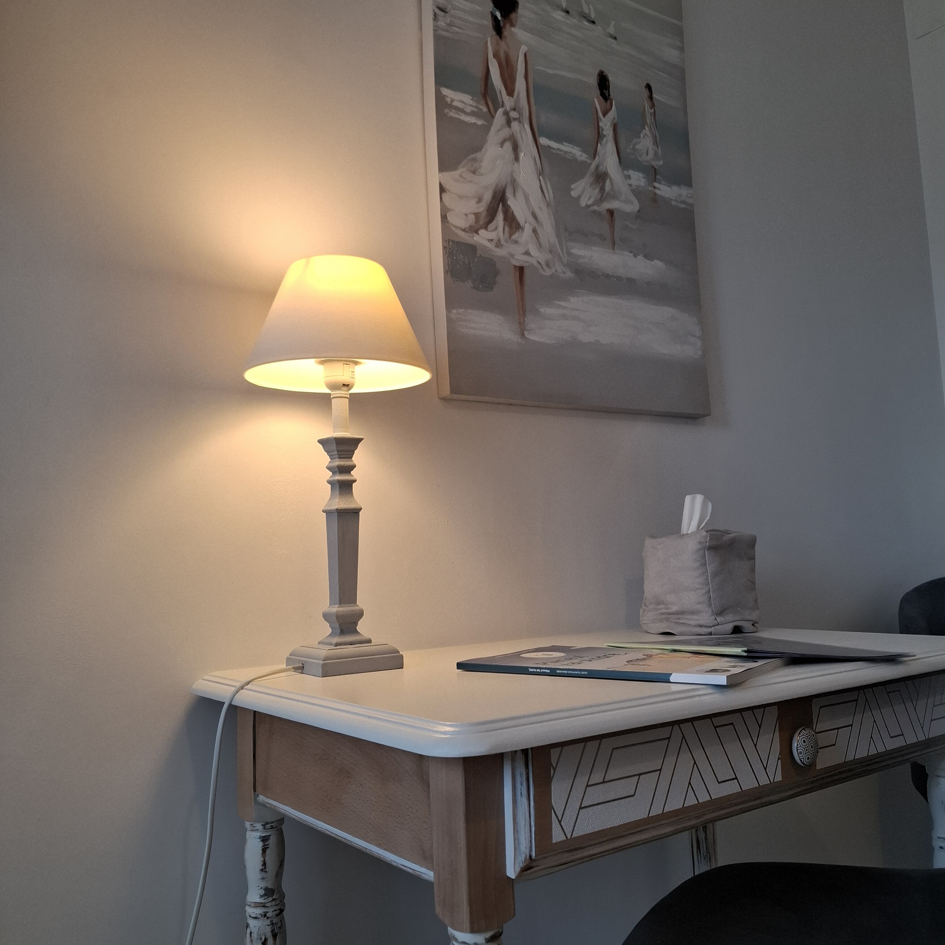 Uma mesa com uma lâmpada, uma caixa de lenços e uma pintura de mulheres em vestidos.