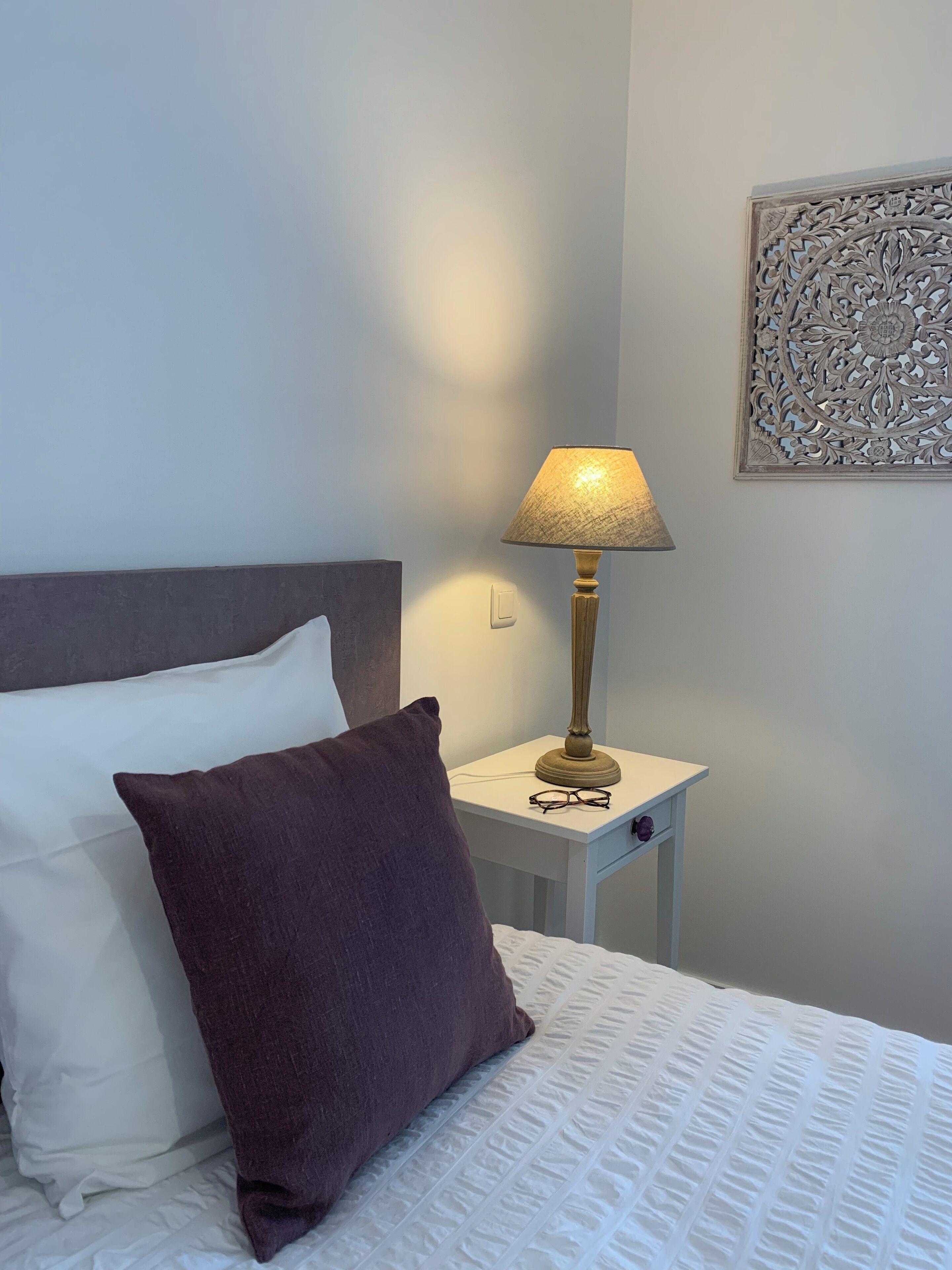 Una cama bien tendida con un cojín blanco y uno morado oscuro, una mesita de noche con una lámpara, y un adorno decorativo en la pared.
