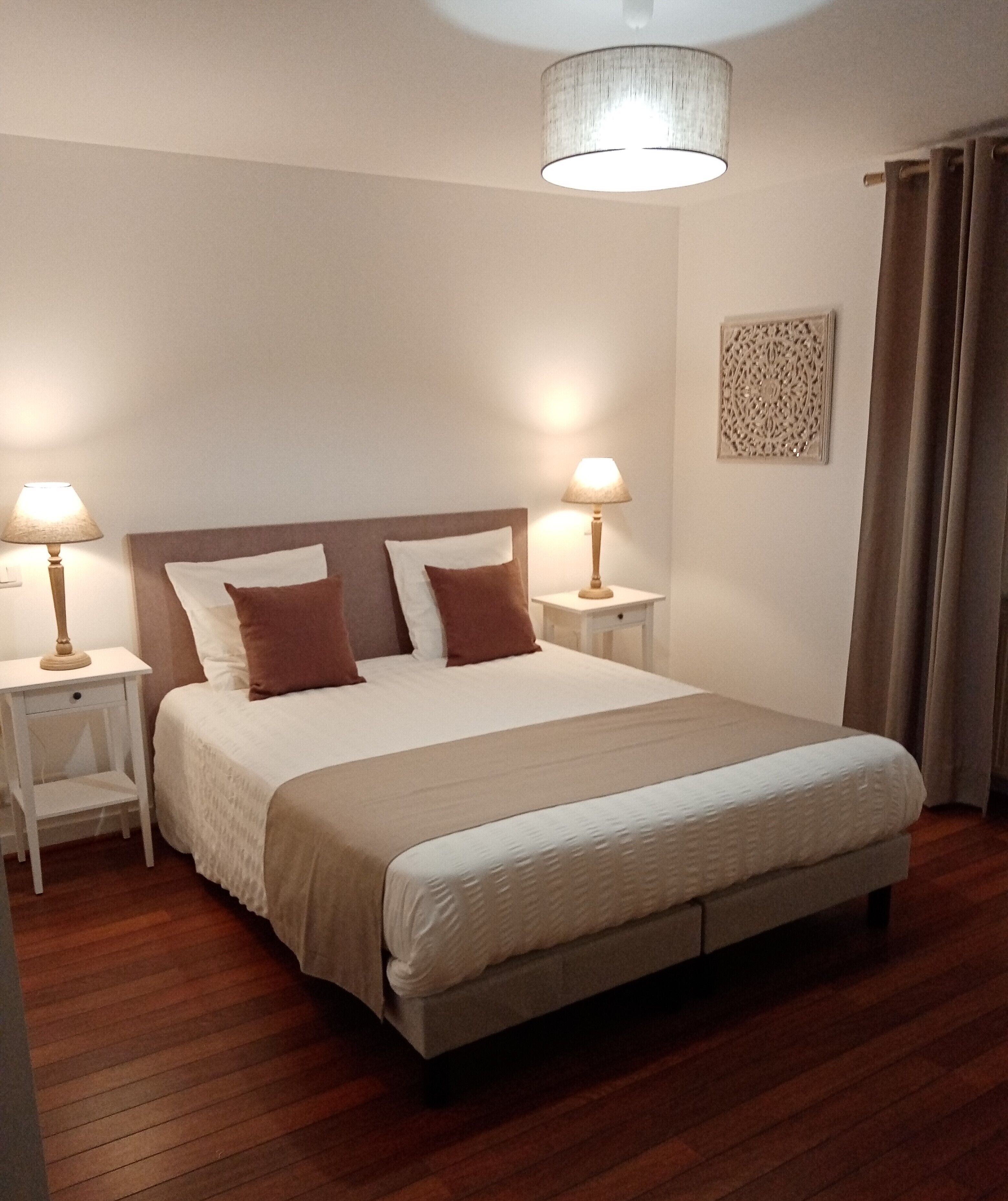 A neatly made bed with a headboard, two bedside tables with lamps, a decorative wall piece, and a window with curtains.