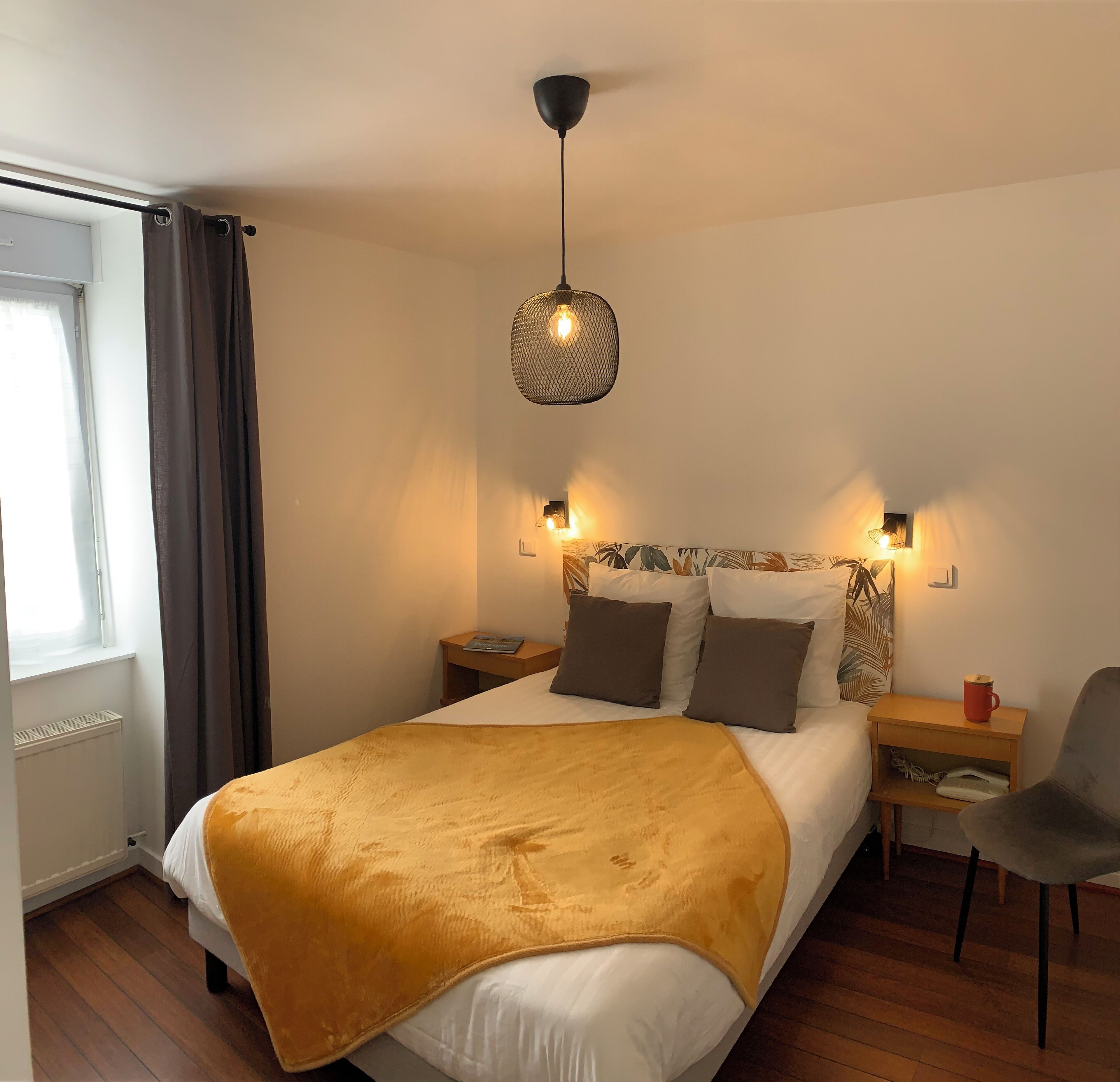 Un dormitorio ordenado con cama, mesitas de noche, una silla y una lámpara colgante.
