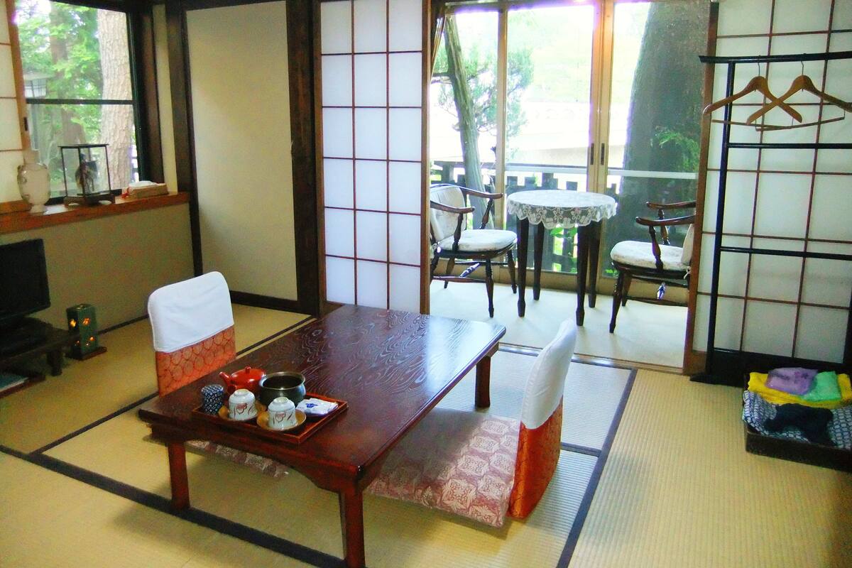 A traditional Japanese room with tatami flooring, a low wooden table with tea set, and sliding doors leading to an outdoor balcony with chairs and a table.