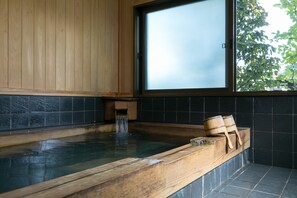 A wooden bathtub with a small waterfall feature, a window with frosted glass, and a view of greenery outside. - Sumiyoshi Ryokan (Takayama)