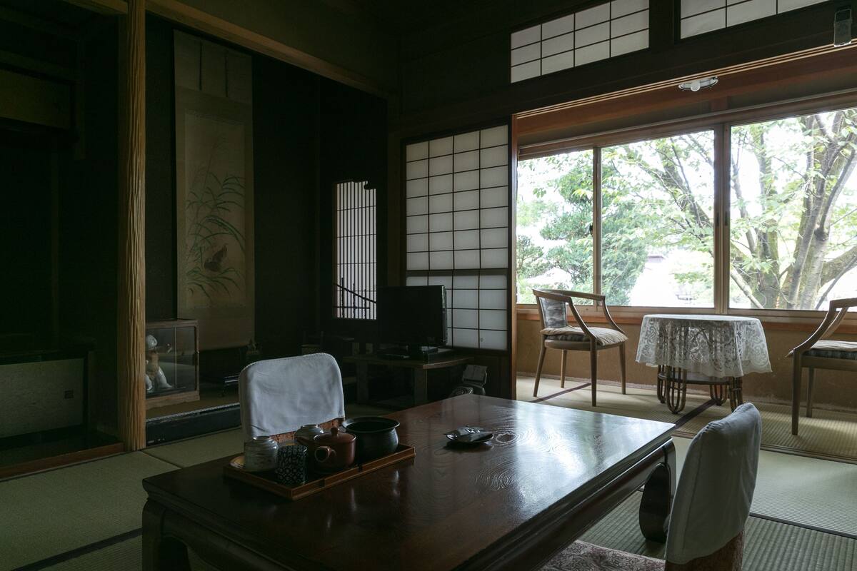 A traditional Japanese room with tatami flooring, a low table with a tea set, a television, and a view of trees through large windows.