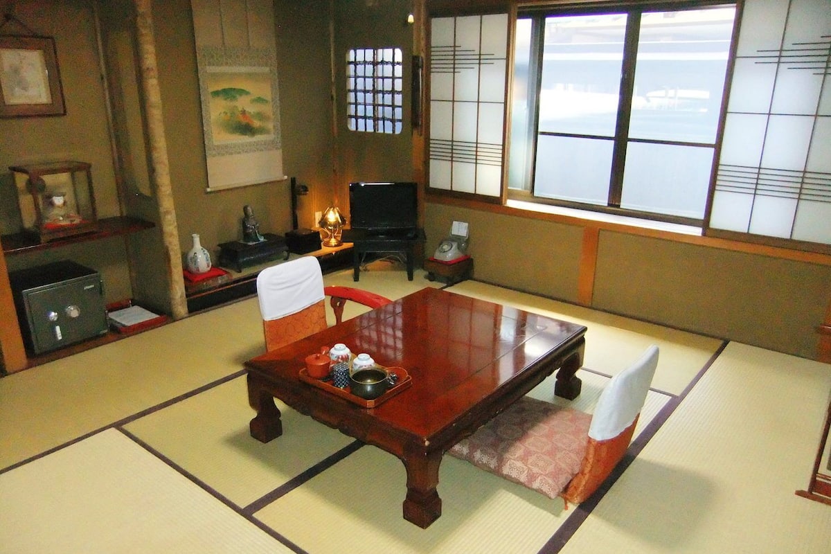 Una habitación japonesa tradicional con suelo de tatami, una mesa baja con un bandeja de utensilios para té, una silla negra y una ventana con vistas al exterior.