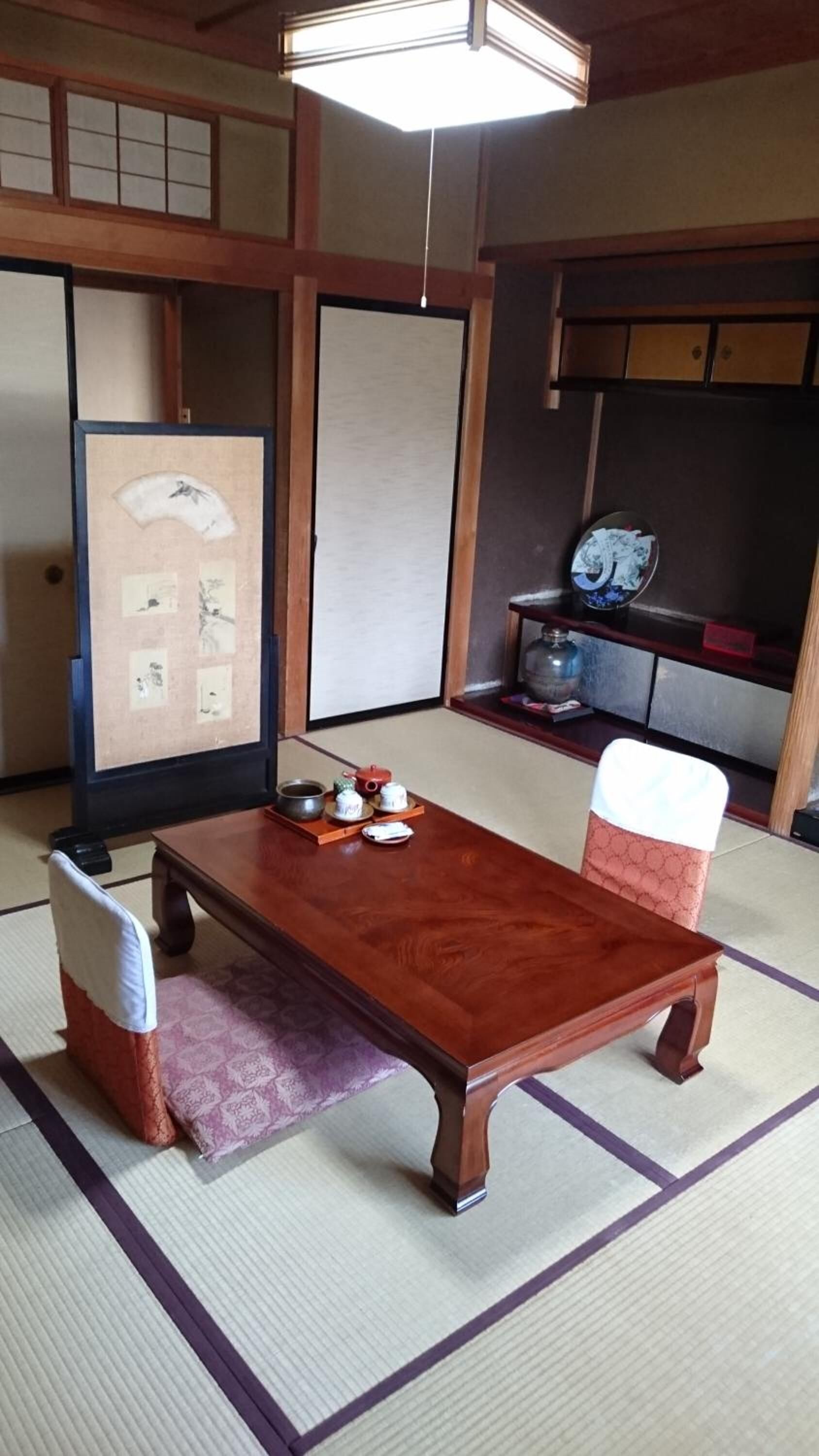 Ett traditionellt japanskt rum med tatamimattor på golvet, ett lågt träbord med teutensilier, en vadderad stol och en skjutdörr med en dekorativ panel.