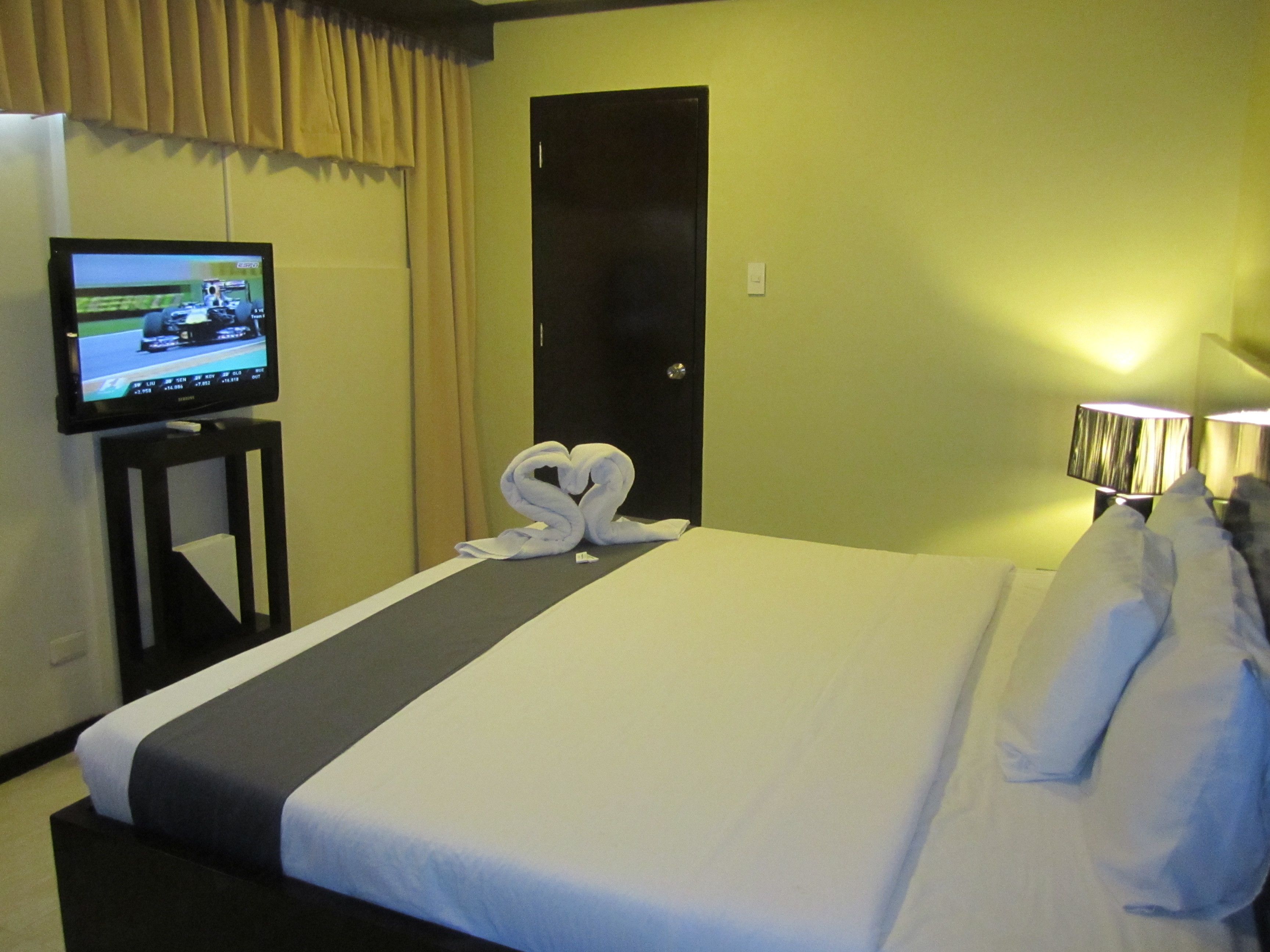 A hotel room with a bed, a television on a stand, a heart-shaped towel on the bed, and a lamp on the nightstand.
