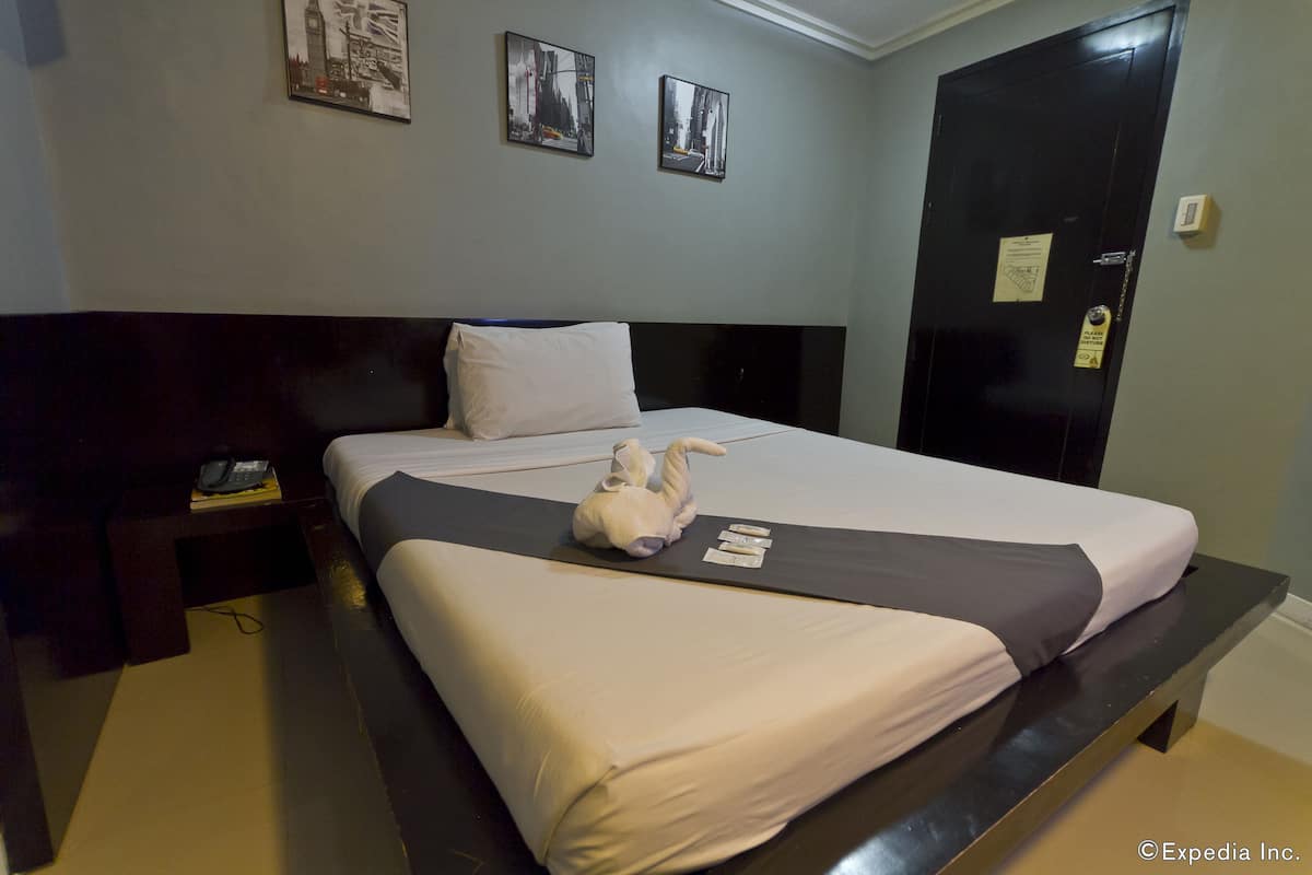A neatly made bed with a white and gray bedspread, a pillow, and a stuffed toy on the bed. There is a nightstand with a phone and a book beside the bed. The room has a dark wooden headboard and a black door with a lock.