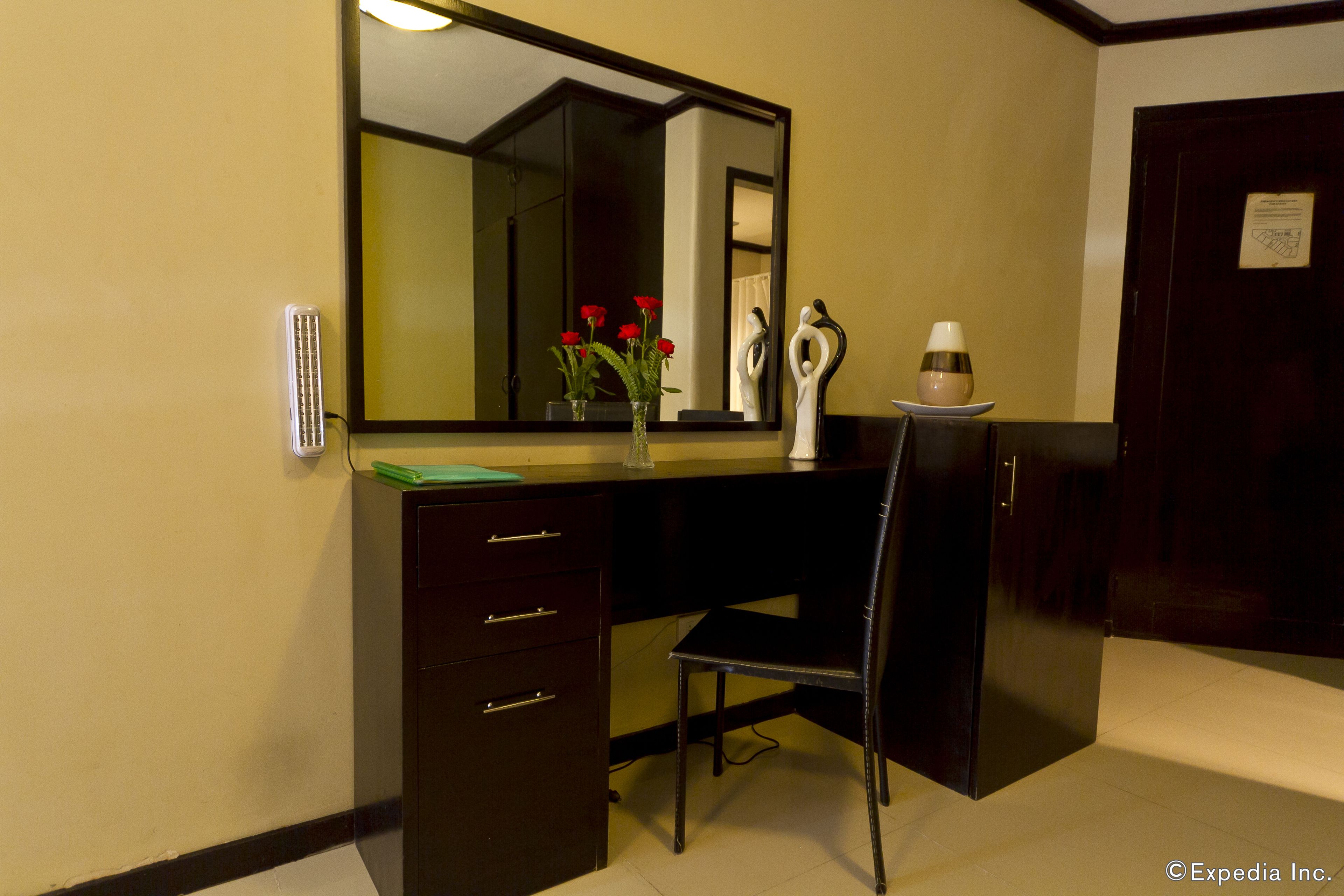 A hotel room with a dark wooden desk, a chair, a mirror, and a vase with red flowers.