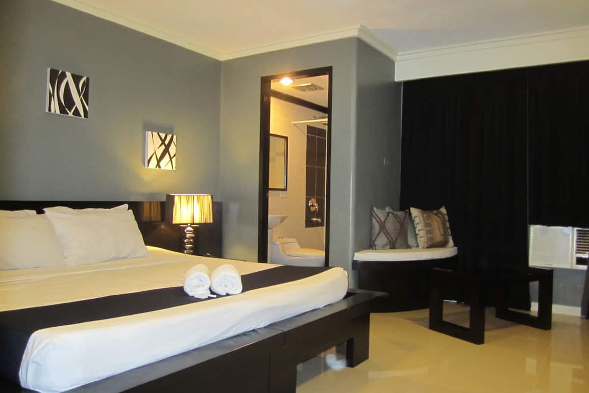 A hotel room with a large bed, a bedside table with a lamp, a chair, and a desk with a computer.