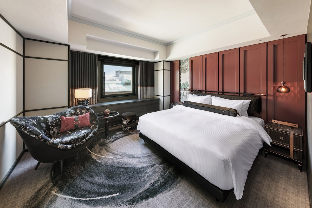 A modern hotel room with a large bed, a chair, a small table, and a view of the city through the window.