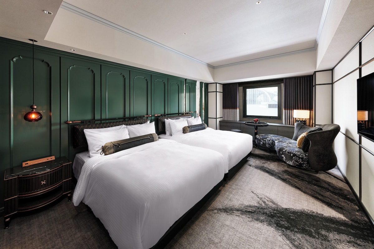 A hotel room with two beds, a desk, a chair, a large window, and a green accent wall.
