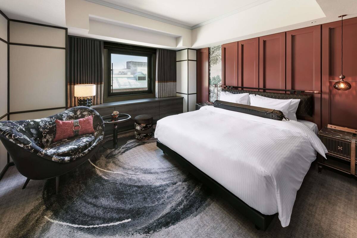 A hotel room with a bed, a sofa, a small table, a lamp, and a window with a view.