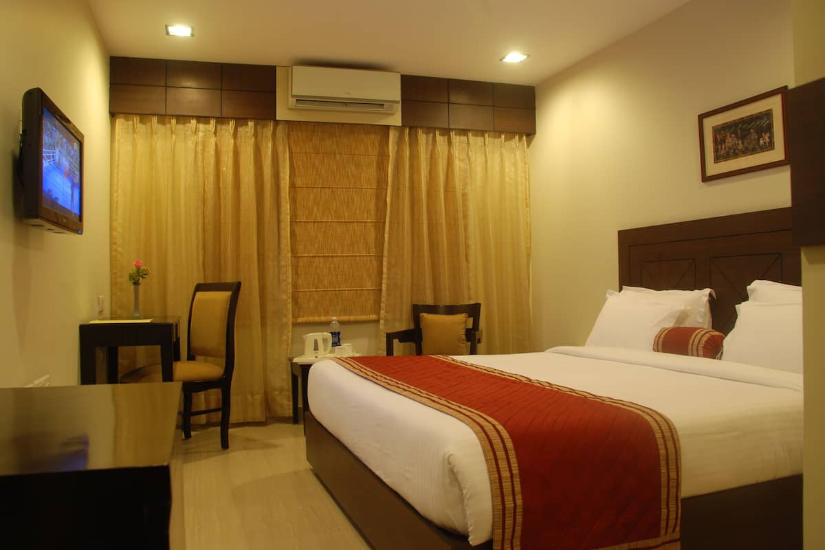 A hotel room with a large bed, a desk, a chair, a small table with a lamp, and an air conditioning unit.