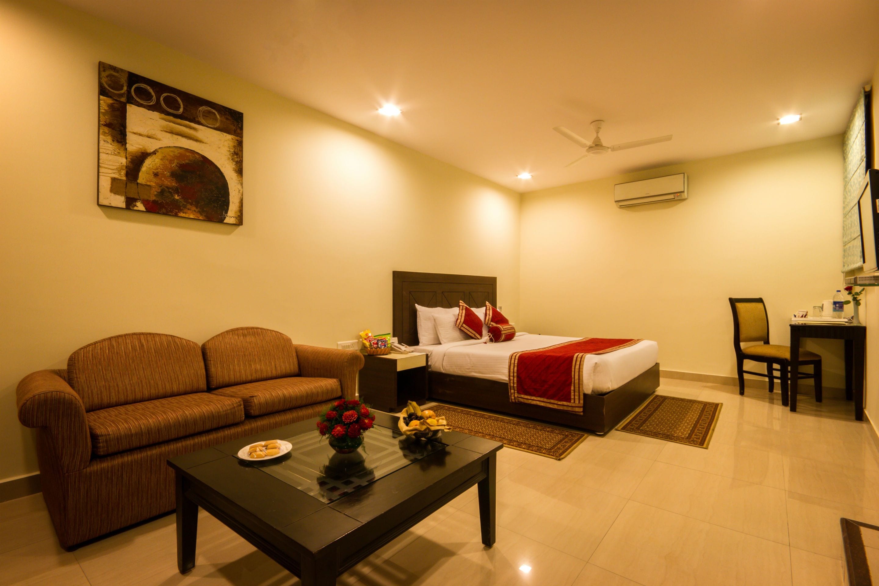 A hotel room with a bed, a sofa, a coffee table with flowers, a desk, and a painting on the wall.
