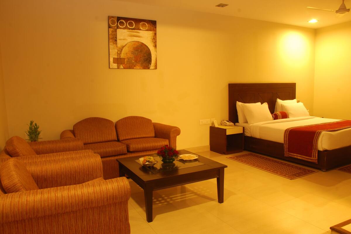 A hotel room with a sofa set, a bed with a red blanket, a nightstand, and a painting on the wall.