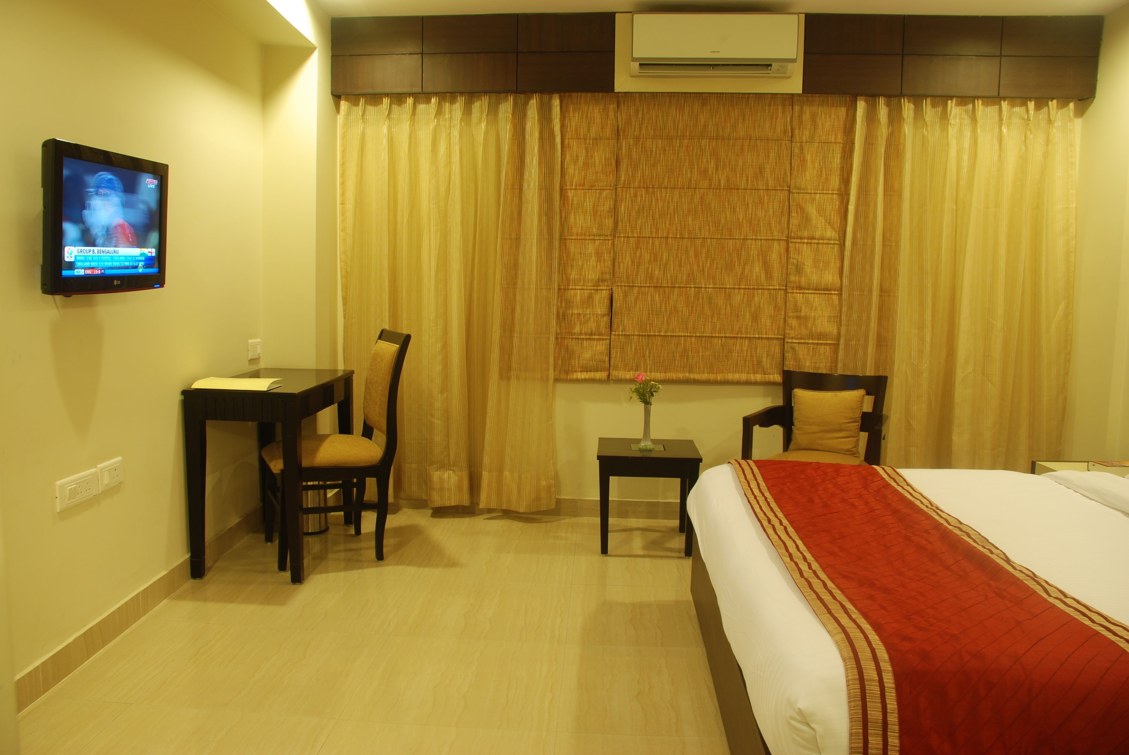 A hotel room with a bed, a desk with a chair, a small table with a vase, and a television on the wall.