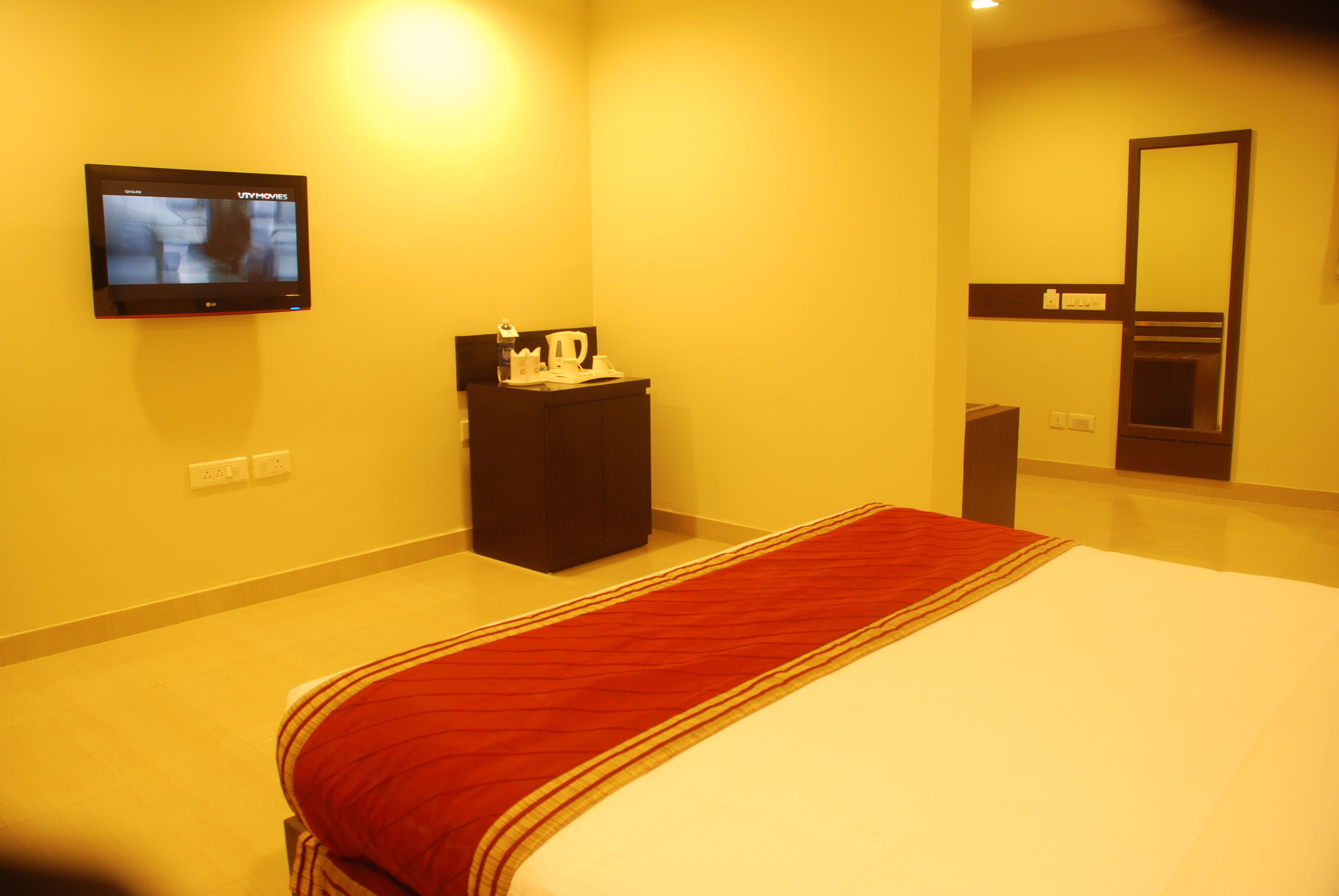 Habitación de hotel con una cama, un escritorio pequeño con un hervidor, un televisor montado en la pared y un espejo sobre el escritorio.