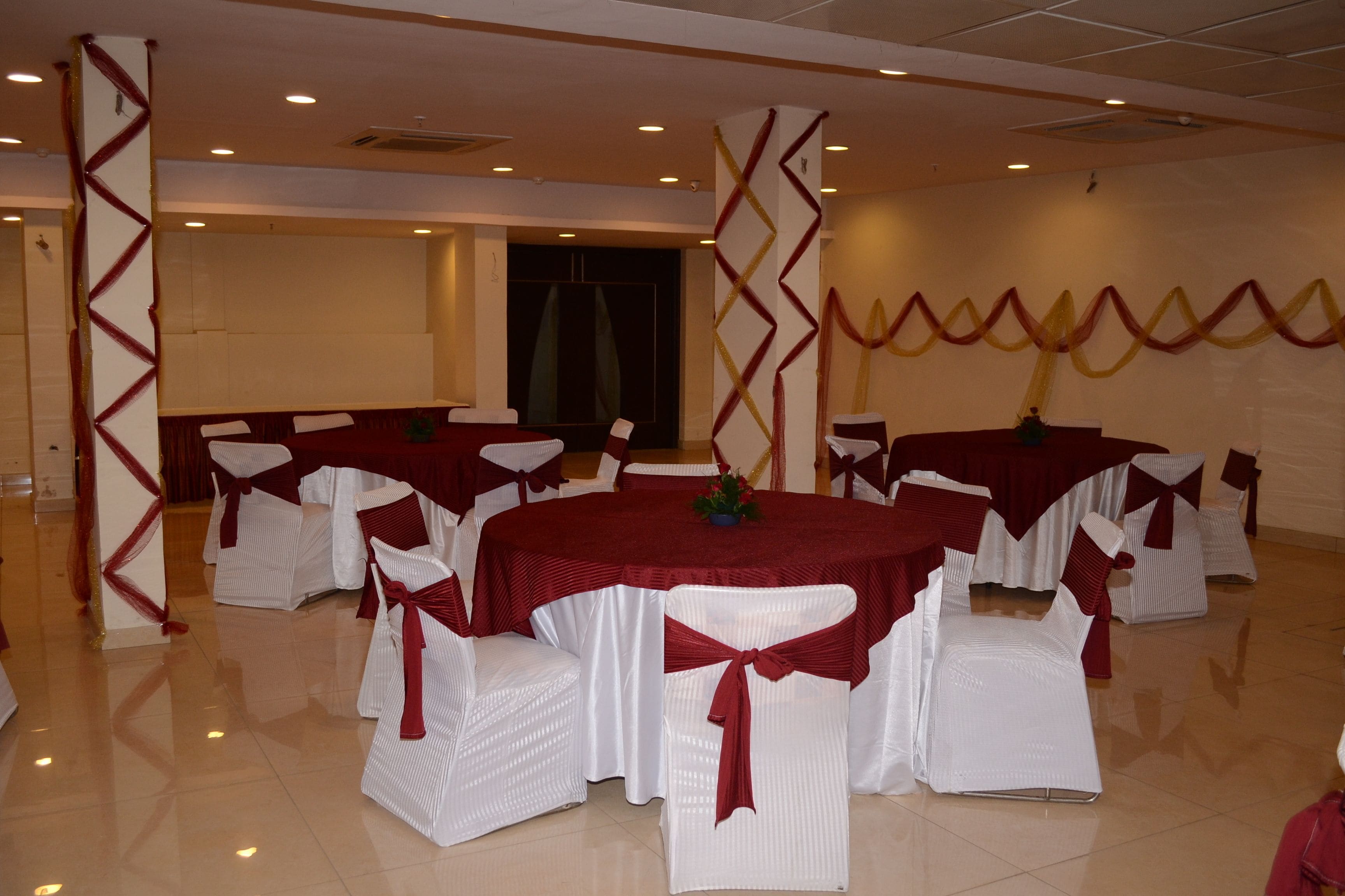 Un salón de banquetes con mesas redondas, sillas blancas adornadas con cintas rojas y un escenario.