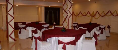 Un salón de banquetes con mesas redondas, sillas blancas adornadas con cintas rojas y un escenario.