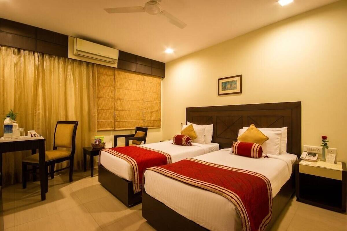 A hotel room with two beds, a desk, a chair, an air conditioner, and a nightstand with a phone and a vase of flowers.