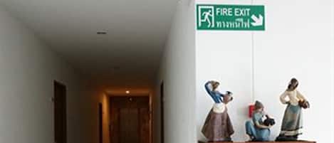 A hotel corridor with a fire exit sign, a red emergency pull station, and a wooden display cabinet with figurines.