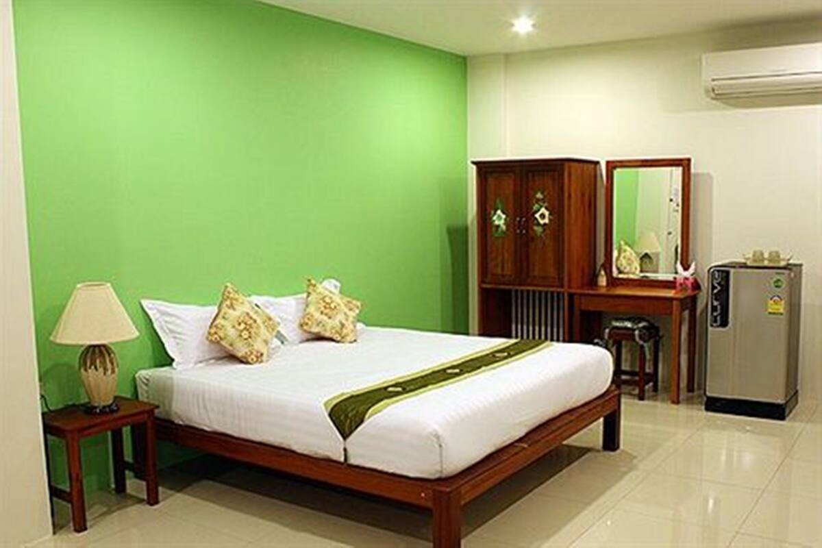 Ein Hotelzimmer mit grüner Wand, einem hölzernen Bett mit weißer Bettwäsche, einem Nachttisch aus Holz mit einer Lampe, einem Holzschrank, einem Spiegel und einem Kühlschrank.
