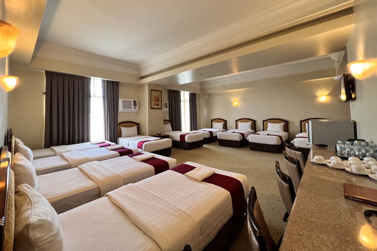 Ein Hotelzimmer mit vier nebeneinander stehenden Betten, einem Schreibtisch mit Stühlen, einer Minibar und einem Fernseher.