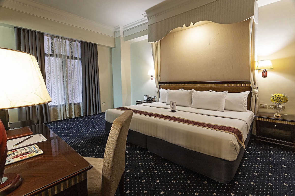 Ein Hotelzimmer mit einem großen Bett, einem Schreibtisch mit Lampe, einem Sessel und einem Nachttisch mit Lampe und Blumen.