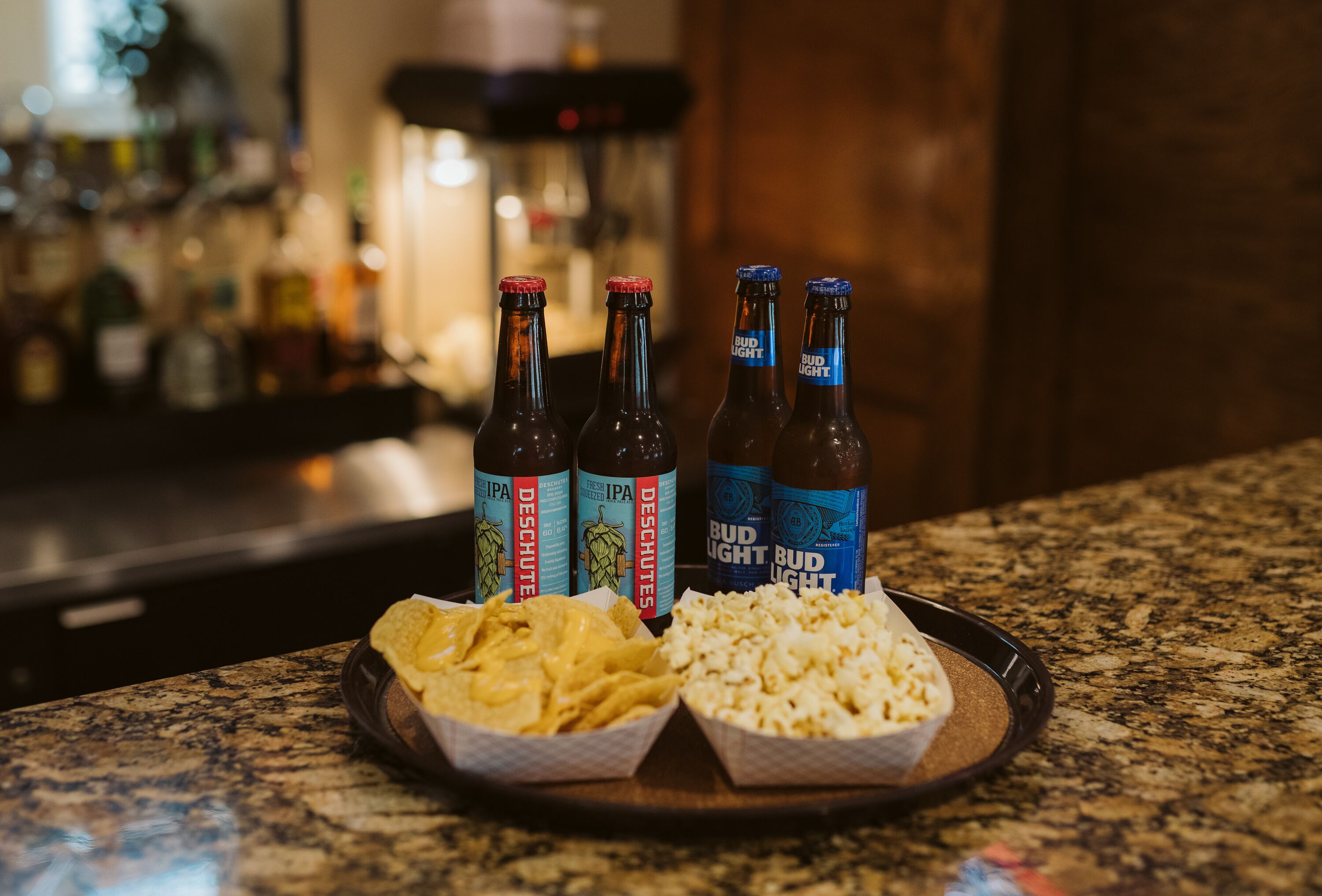 A tray with nachos and popcorn on a granite countertop, accompanied by four bottles of Bud Light and Deschutes IPA.