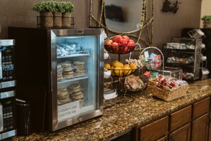 A self-service breakfast station with a refrigerated display case, baskets of fresh fruits, and a variety of packaged goods. - EverSpring Suites (Bismarck)