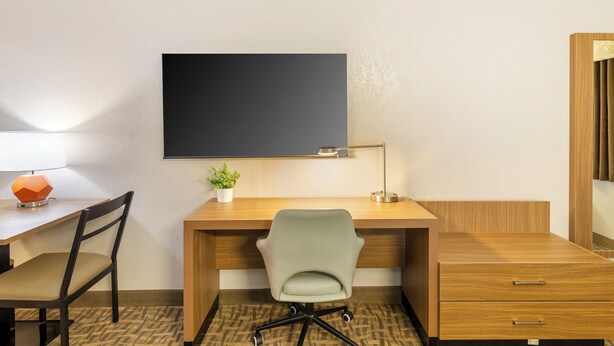 A hotel room with a wooden desk, a green office chair, a flat-screen TV mounted on the wall, a small potted plant, and a desk lamp. - EverSpring Suites (Bismarck)