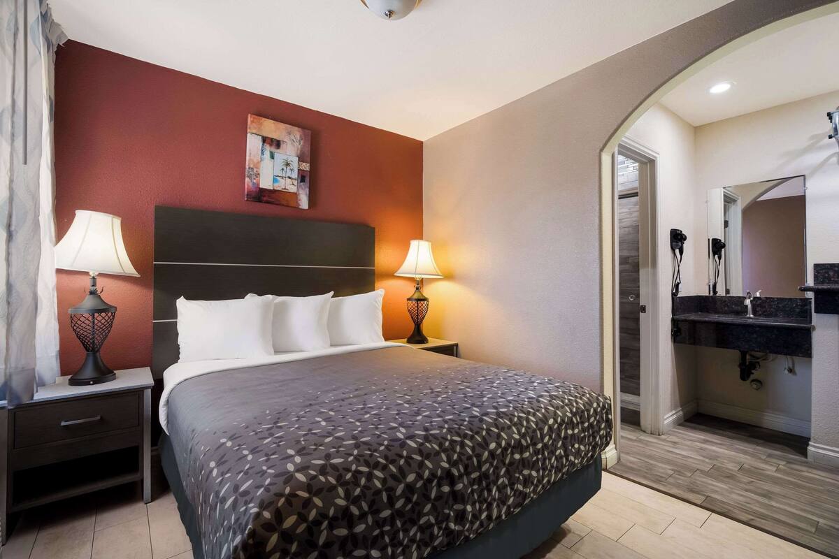 A hotel room with a bed, two bedside lamps, a nightstand, a painting on the wall, and a bathroom visible through an archway.