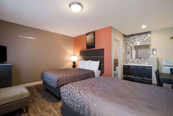 A hotel room with two beds, a desk, a chair, a TV, and a kitchenette.