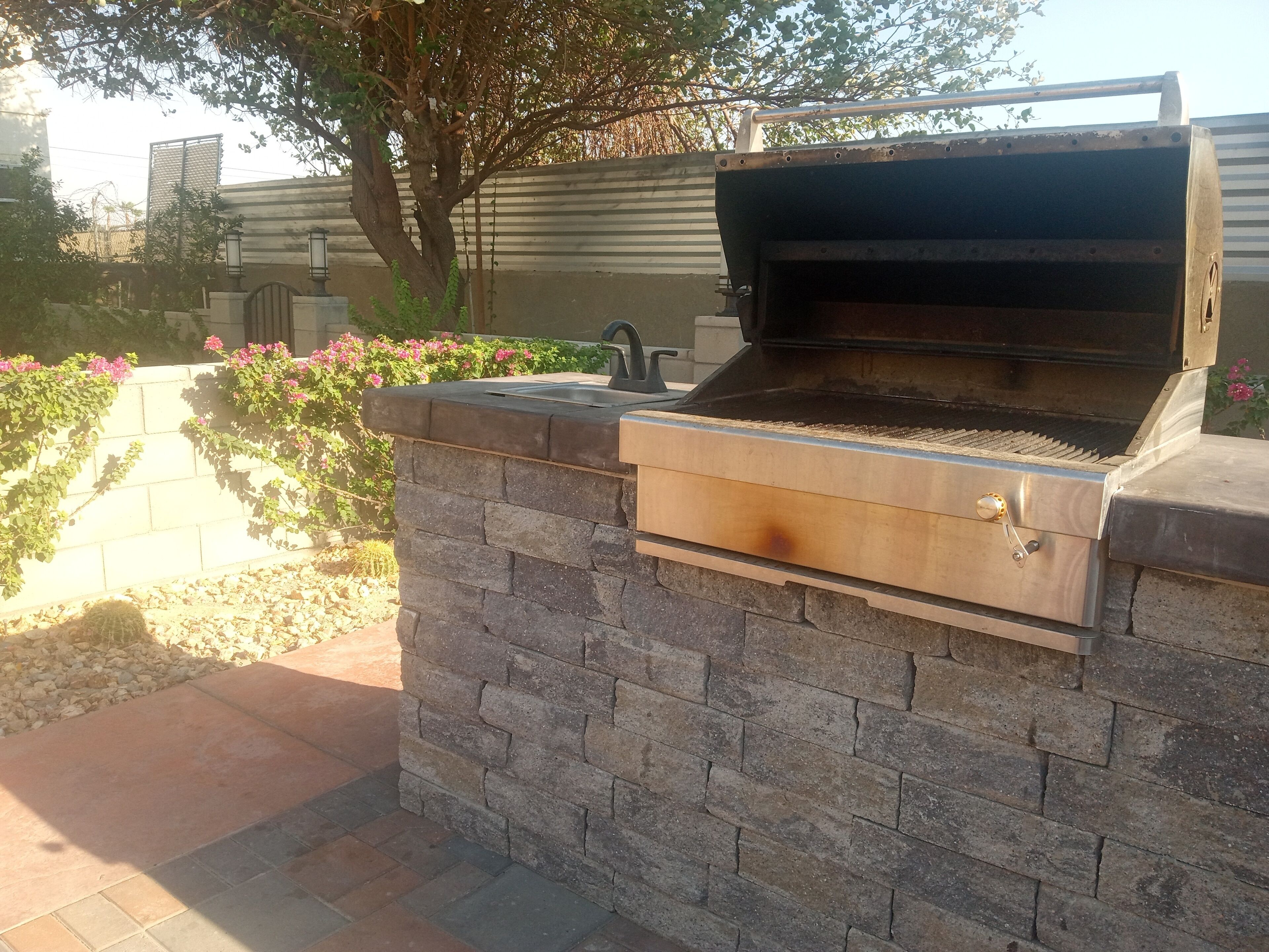 An outdoor grill with a stone base and a metal lid, set against a backdrop of a garden with flowering plants and a tree.