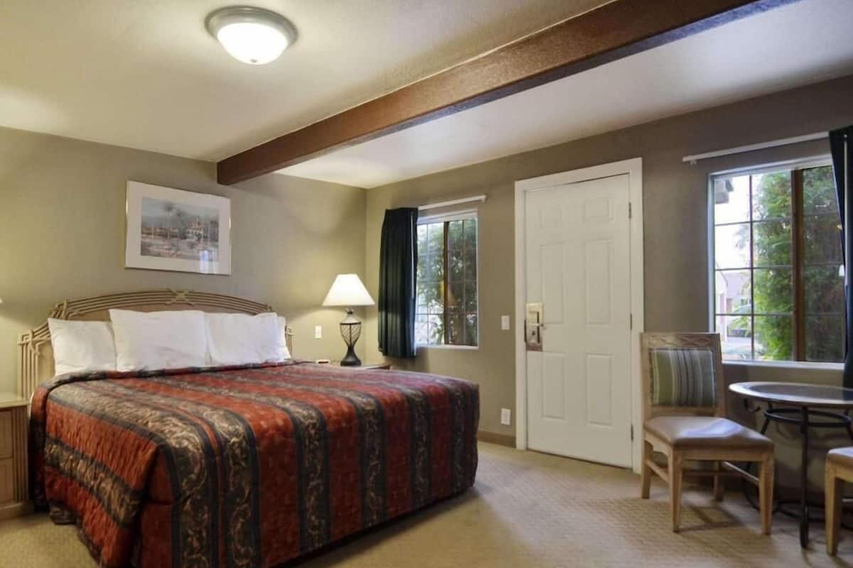 A hotel room with a bed, a chair, a table, a lamp, a door, and a window.