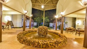 A well-lit indoor area with a central water feature surrounded by stone, wooden pillars, and seating areas. - Kena Beach Hotel (Matemwe)