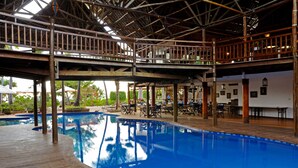 An indoor swimming pool with wooden deck and lounge area, surrounded by a covered deck with seating and a view of greenery outside. - Kena Beach Hotel (Matemwe)
