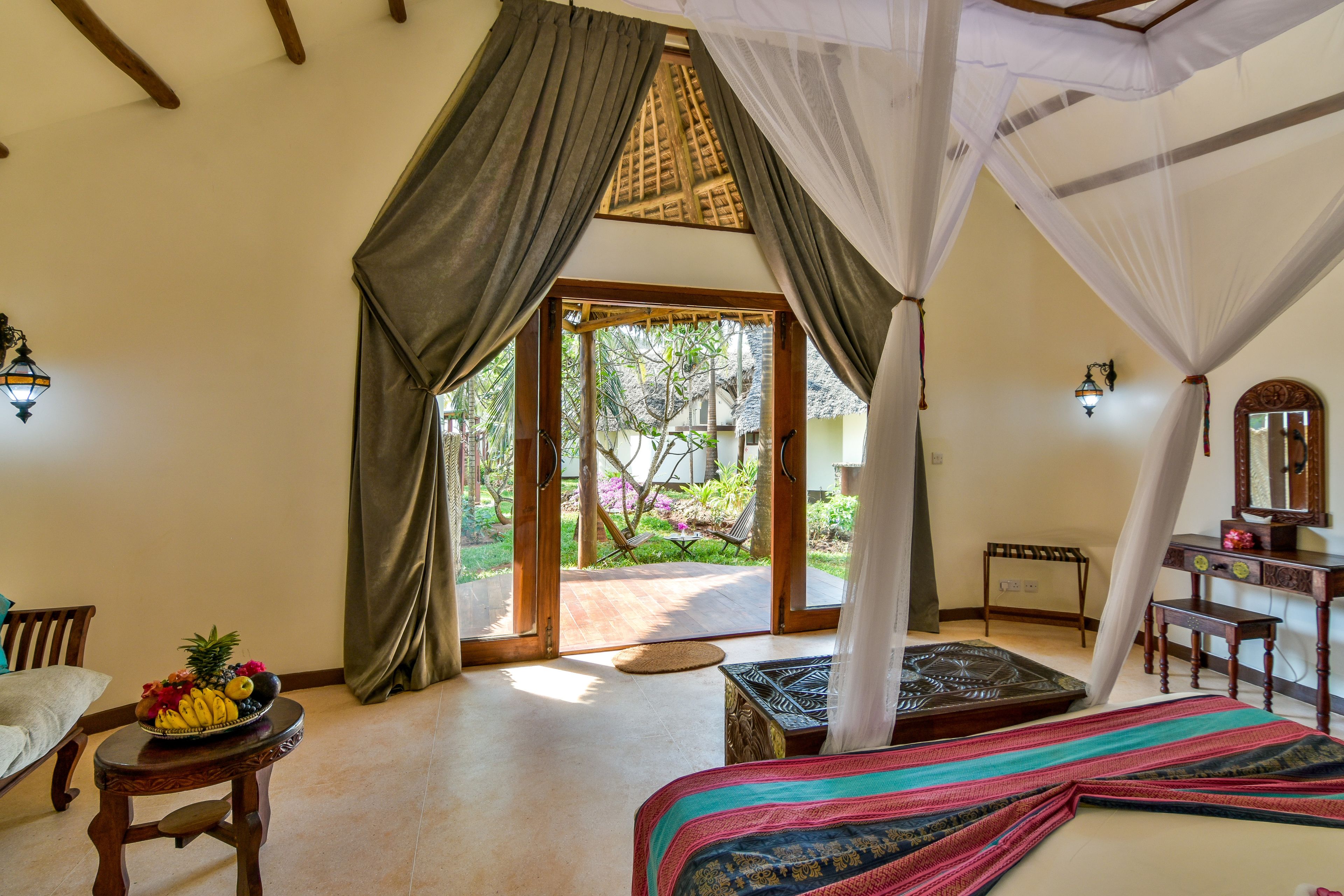 Ein Zimmer mit einem Bett, einem kleinen Tisch mit Obst, einem Stuhl, einer hölzernen Bank und Blick ins Freie.