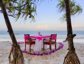 Une table installée sur la plage, recouverte d’une nappe blanche et agrémentée de décorations roses, entourée de chaises, avec en arrière-plan des palmiers et la mer.